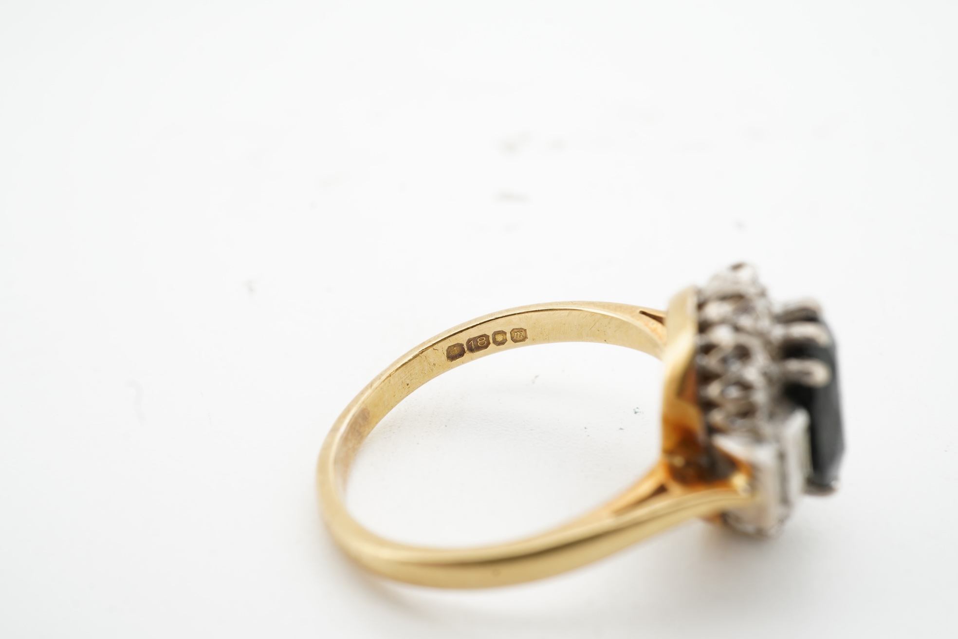 A sapphire and diamond cluster ring, claw-set with an oval sapphire measuring approximately 8.2 x 6.1 x 4.3mm, within a border of brilliant-cut and baguette diamonds totalling approximately 0.20 carats, mounted in 18ct g